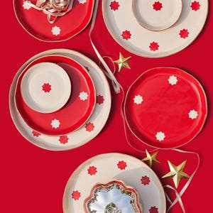 May include: A collection of round ceramic plates and bowls in red and cream, featuring repeating star-shaped patterns. The set includes a silver-toned metallic pumpkin-shaped dish, gold star ornaments, and a light-coloured ribbon.