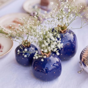 May include: Three deep blue ceramic pomegranate-shaped vases with metallic gold-coloured rims and small gold dots scattered across the surface. Each vase contains a delicate arrangement of white baby's breath flowers with thin green stems.