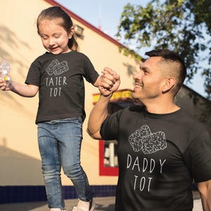 May include: Matching black t-shirts with white text and a tater tot graphic. One shirt reads "TATER TOT," the other reads "DADDY TOT." A child and an adult are holding hands, wearing the shirts.