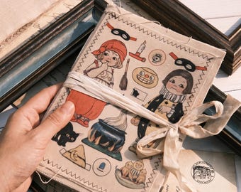 Puede incluir: Una cubierta de tela rectangular para diario hecha de lona color crema con un patrón impreso de dos niños, un gato negro, un caldero, una escoba y máscaras. Los bordes tienen una costura en zigzag y está atada con una cinta blanca.