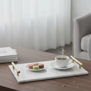 May include: A rectangular white marble serving tray featuring two gold-toned metal handles on the sides. The tray holds a small white saucer with four colorful macarons and a white ceramic teacup filled with a steaming hot beverage.