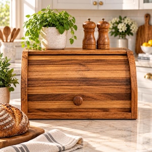 May include: A rectangular wooden bread box with a roll-top lid and a central wooden knob. The box features horizontal slats of varying brown wood tones. Two wooden salt and pepper grinders sit on top of the bread box next to a potted plant.