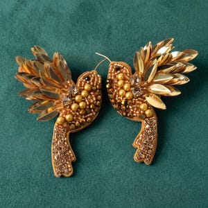 May include: A pair of bird-shaped brooches crafted with gold-toned sequins, metallic beads, and small crystals. Each bird features layered sequin wings, a beaded body with round golden pearls, and a tail embellished with sparkling brown crystals.
