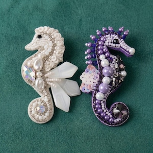 May include: Two seahorse-shaped brooches crafted with intricate beadwork. The left brooch features white beads, pearls, and iridescent crystals. The right brooch displays purple beads, pearls, and dark faceted crystals with light purple sequins.
