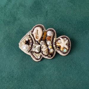 Puede incluir: Un broche con forma de pez que presenta una concha de cauri, una pequeña estrella de mar y varias cuentas blancas y marrones. El diseño está delineado con hilo marrón oscuro y decorado con cristales transparentes, perlas y elementos metálicos.
