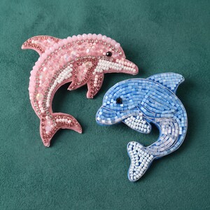 May include: Two dolphin-shaped brooches crafted with intricate beadwork. One brooch features light pink and iridescent beads with a dark eye, while the other is composed of various shades of blue and white beads with a dark eye, both in a leaping pose.