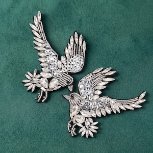May include: Two bird-shaped patches crafted with silver sequins, clear faceted crystals, and metallic beads. Each bird is depicted in flight with wings extended, showcasing intricate embroidery and reflective embellishments on a dark base.