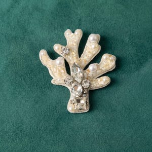 Op de afbeelding: Een koraalvormige broche met een crèmekleurige basis gevuld met kleine, getextureerde kralen. Het sieraad is versierd met diverse heldere, gefacetteerde kristallen en ronde, witte imitatieparels die over de takken zijn verdeeld.