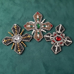 Puede incluir: Tres parches decorativos en forma de cruz adornados con cuentas, pedrería y cristales. El parche izquierdo tiene cuentas doradas, el central piedras verdes y rojas, y el derecho muestra una gran piedra central de color rojo.