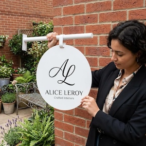 Puede incluir: Un letrero circular blanco con un logotipo negro que presenta las letras estilizadas AL, el texto ALICE LEROY y las palabras Crafted Interiors debajo. El letrero está sujeto a un soporte de montaje horizontal de metal blanco.
