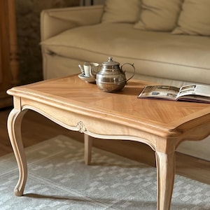 Puede incluir: Una mesa de centro rectangular de madera con acabado marrón claro, que presenta un patrón de espiga en el tablero, patas curvas tipo cabriolé y detalles florales tallados en el faldón. Una tetera de plata y un libro abierto descansan sobre ella.