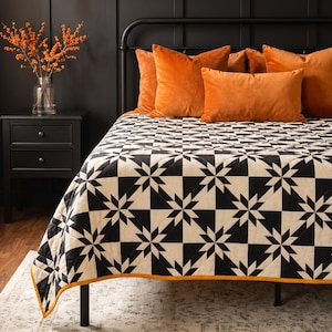 May include: A black and white quilt featuring a repeating star pattern with a thin orange border. The quilt is draped over a black metal bed frame adorned with several solid orange velvet throw pillows.