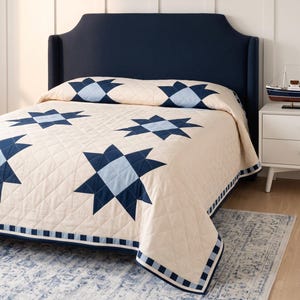 May include: A cream-colored quilt featuring a repeating star pattern in navy blue and light blue. The quilt has a diamond-stitched texture and a border with a navy and light blue checkered design, draped over a bed with a navy blue headboard.