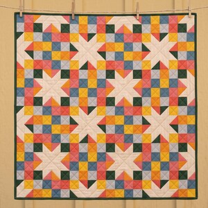 May include: A square quilt featuring a repeating star pattern composed of small fabric squares in shades of cream, yellow, pink, blue, and dark green. The quilt is finished with a dark green border and displays visible grid-like stitching.