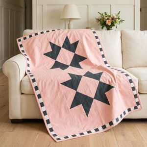 May include: A light pink quilt featuring two large dark charcoal gray star-shaped patterns centered on the fabric. The quilt is finished with a decorative border consisting of alternating small black and white square blocks along the edges.