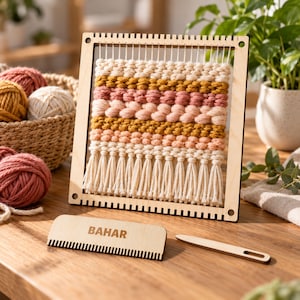 May include: A square wooden weaving loom frame holding a partially completed tapestry with horizontal rows of cream, mustard yellow, and dusty rose yarn, finished with a fringe at the bottom. A wooden weaving comb labelled BAHAR and a shuttle.