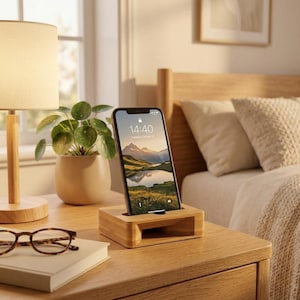 May include: A light-colored wooden smartphone stand with a rectangular base and a horizontal slot for device placement. The stand features a hollow acoustic chamber design and is positioned on a wooden surface next to a book and eyeglasses.