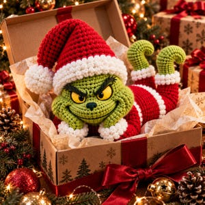 May include: A crocheted green creature with yellow eyes and a grumpy expression, wearing a red and white Santa hat and matching red and white suit, resting inside an open cardboard gift box decorated with a pattern of pine trees and snowflakes.