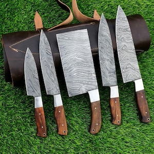 May include: A set of five kitchen knives with Damascus steel blades featuring a wavy pattern and dark brown wooden handles with silver rivets, arranged in front of a dark brown leather knife roll with a strap and buckle closure.