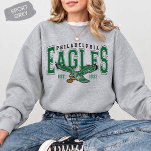 May include: A sport grey crewneck sweatshirt featuring the text Philadelphia Eagles in green with white outlines, an eagle graphic holding a football, and the text EST 1933 in green. A grey tag in the corner reads SPORT GREY.