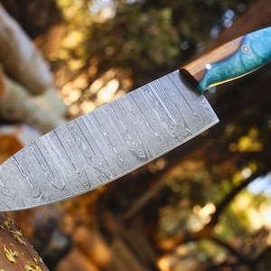 May include: A chef knife with a patterned Damascus steel blade and a handle crafted from a combination of natural brown wood and a translucent teal resin, featuring a small circular metal inlay on the side of the handle near the bolster.