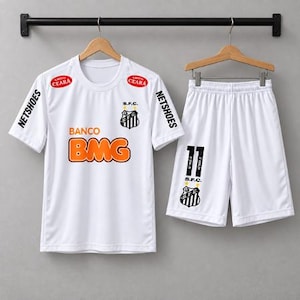Può includere: Un completo da calcio bianco composto da maglia e pantaloncini, con lo stemma S.F.C., la scritta arancione BANCO BMG, NETSHOES sulle maniche e i loghi ALIMENTOS CEARA. I pantaloncini riportano il numero 11, lo stemma S.F.C. e NEYMAR JR.