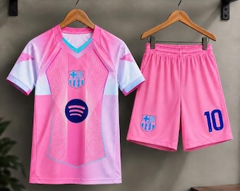 Könnte beinhalten: Ein Fußballtrikot-Set in Pink und Hellblau, bestehend aus einem Trikot und passender Hose. Das Trikot hat einen V-Ausschnitt, das FC Barcelona-Wappen und ein dunkelblaues Spotify-Logo auf der Brust. Die Hose zeigt das FC Barcelona-Wappen und die Nummer 10.