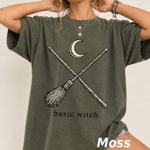 Può includere: Una t-shirt a maniche corte color verde muschio con una stampa grafica di una luna crescente e due scope incrociate. Sotto la grafica, il testo "basic witch" è stampato in un carattere serif nero. La maglietta ha un girocollo.