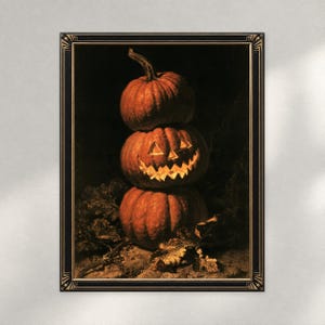 May include: A framed wall art print featuring a stack of three orange pumpkins against a dark background. The middle pumpkin is carved with a glowing jack-o'-lantern face, while the top and bottom pumpkins remain whole. The frame is black with gold.