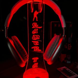 Könnte beinhalten: Ein weißer Over-Ear-Kopfhörer auf einem transparenten Acrylständer, der in hellem Rot leuchtet. Der Ständer zeigt eine eingravierte Soldaten-Silhouette und den vertikal angeordneten Schriftzug "GAMER TAG" in einer stilisierten Schriftart.