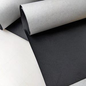 May include: Two rolls of black fabric with a fine, textured weave. Each roll features a white, semi-transparent protective paper backing that is partially peeled away to reveal the solid black material underneath, showing a consistent matte finish.