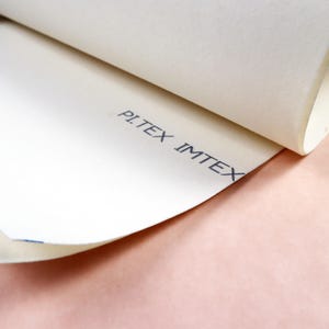 Può includere: Una pila di fogli di materiale spessi color bianco sporco con una consistenza liscia e opaca. Il foglio superiore è parzialmente ripiegato, rivelando il testo PI.TEX IMTEX stampato in inchiostro nero sulla superficie del foglio sottostante.