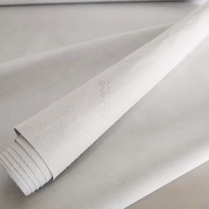 Può includere: Un rotolo di pelle scamosciata grigio chiaro con una consistenza morbida e opaca. La superficie della pelle presenta il testo in rilievo "CYLIANE" seguito da un testo più piccolo che recita "HANDCRAFTED" in un carattere sans-serif pulito.