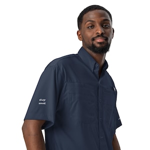 May include: A navy blue short-sleeved button-down shirt featuring a pointed collar, two front chest pockets, and the text "chop wood" printed in white script on the right sleeve. The shirt is made of a smooth, lightweight fabric.