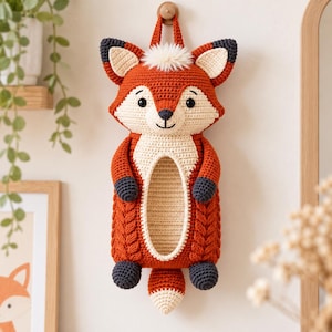 May include: A hanging crochet fox wall organizer featuring a rust-orange and cream color palette. The fox has pointed ears, a small tuft of white faux fur on its head, dark gray paws, and a central oval opening for storage with a cable-knit texture.