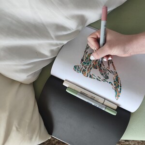 May include: An open sketchbook with a white page featuring a colorful, intricate abstract drawing being colored with a pink marker. Two additional markers in light green and beige are resting on the black cover of the sketchbook.