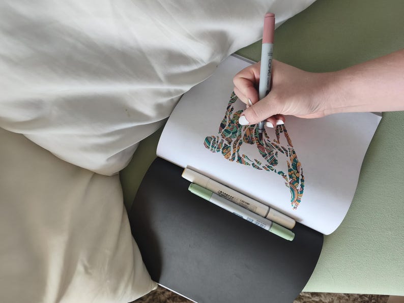 May include: An open sketchbook with a white page featuring a colorful, intricate abstract drawing being colored with a pink marker. Two additional markers in light green and beige are resting on the black cover of the sketchbook.