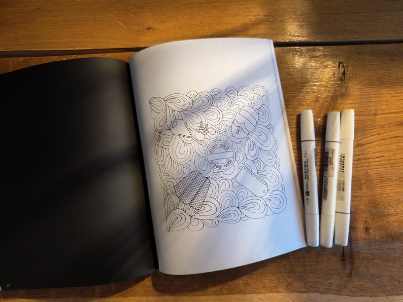 May include: An open coloring book with a black page on the left and a white page on the right featuring a line art illustration of abstract swirls and various objects, accompanied by three white markers with black text on their barrels.