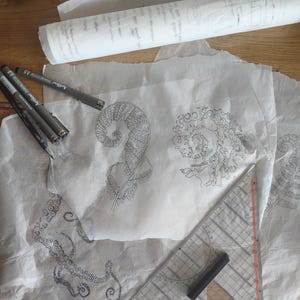 May include: Several sheets of translucent white tracing paper featuring intricate black ink illustrations of a fern frond and an octopus tentacle. Three black fine-liner pens and a clear plastic triangular ruler are placed on the paper.