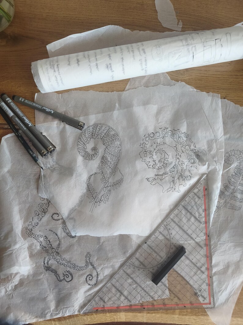 May include: Several sheets of translucent white tracing paper featuring intricate black ink illustrations of a fern frond and an octopus tentacle. Three black fine-liner pens and a clear plastic triangular ruler are placed on the paper.