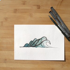 May include: A white rectangular piece of paper featuring a hand-drawn illustration of stylized ocean waves in shades of teal and black ink. Three black pens are positioned diagonally across the top right corner of the paper.