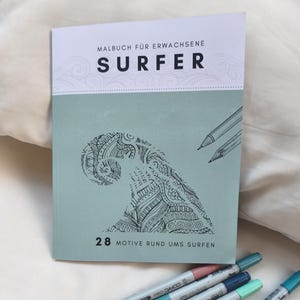 May include: A softcover coloring book titled Malbuch f&uuml;r Erwachsene Surfer with a sage green cover featuring a black line art illustration of a stylized ocean wave. The cover includes the text 28 Motive rund ums Surfen and two pencil sketches.