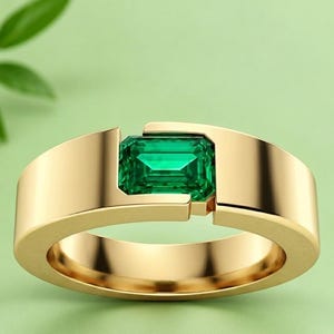 May include: A gold ring featuring a single emerald-cut green gemstone set horizontally within a wide, smooth, polished gold band. The metal band has a distinct open-gap design where the rectangular green stone is securely held in place.