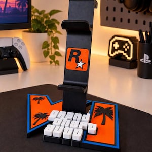 May include: A black vertical gaming headset stand featuring an orange Rockstar Games logo on the front. The base is shaped like the Grand Theft Auto logo with orange and blue accents and white 3D lettering that reads Grand Theft Auto.