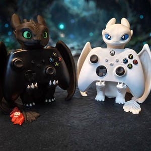 Peut inclure: Deux supports de manette en forme de dragon, l'un noir aux yeux verts et l'autre blanc aux yeux bleus, tenant chacun une manette de jeu assortie avec des boutons colorés, incluant le logo Xbox et les commandes directionnelles standard.