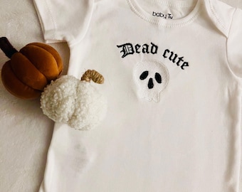 Może przedstawiać: Białe body z krótkim rękawem z wyhaftowanym czarnym napisem Dead cute nad grafiką białej czaszki z czarnymi oczodołami i otworem nosowym. Obok body znajdują się dwie małe ozdobne dynie, jedna brązowa i jedna biała.