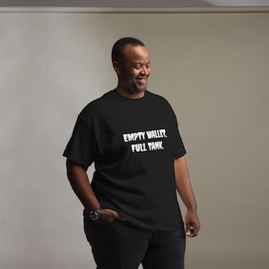 Może przedstawiać: Czarny t-shirt z krótkim rękawem z białym napisem "EMPTY WALLET, FULL TANK." nadrukowanym pogrubioną, postarzaną czcionką na klatce piersiowej. Odzież posiada okrągły dekolt i standardowy krój, zapewniając prosty i schludny wygląd.