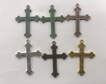 May include: Six decorative crosses in various metallic finishes: teal, green, copper, silver, bronze, and gold. Each cross features an ornate, symmetrical design with a loop at the top for hanging. The crosses are arranged on a light-colored surface.
