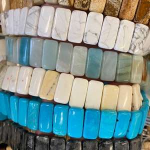May include: A stack of rectangular gemstone beads in various colors, including white, light blue, beige, turquoise, and black. The beads are arranged in rows, with each row featuring a different color.