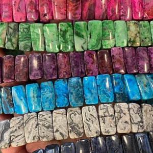 May include: A collection of colorful agate gemstone beads in rectangular shapes. The beads are arranged in rows, showcasing various colors and patterns, including pink, green, purple, blue, and gray.